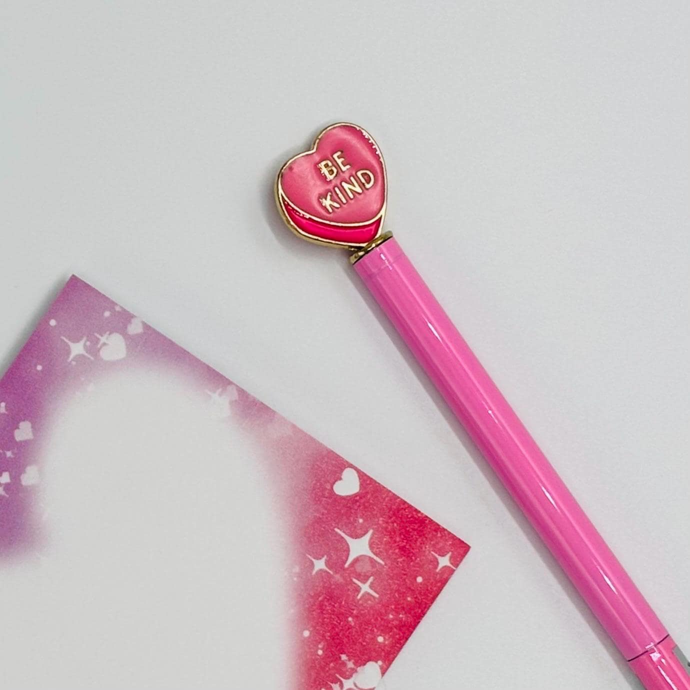 Pink pen with heart-shaped 'Be Kind' clip on a light gray background